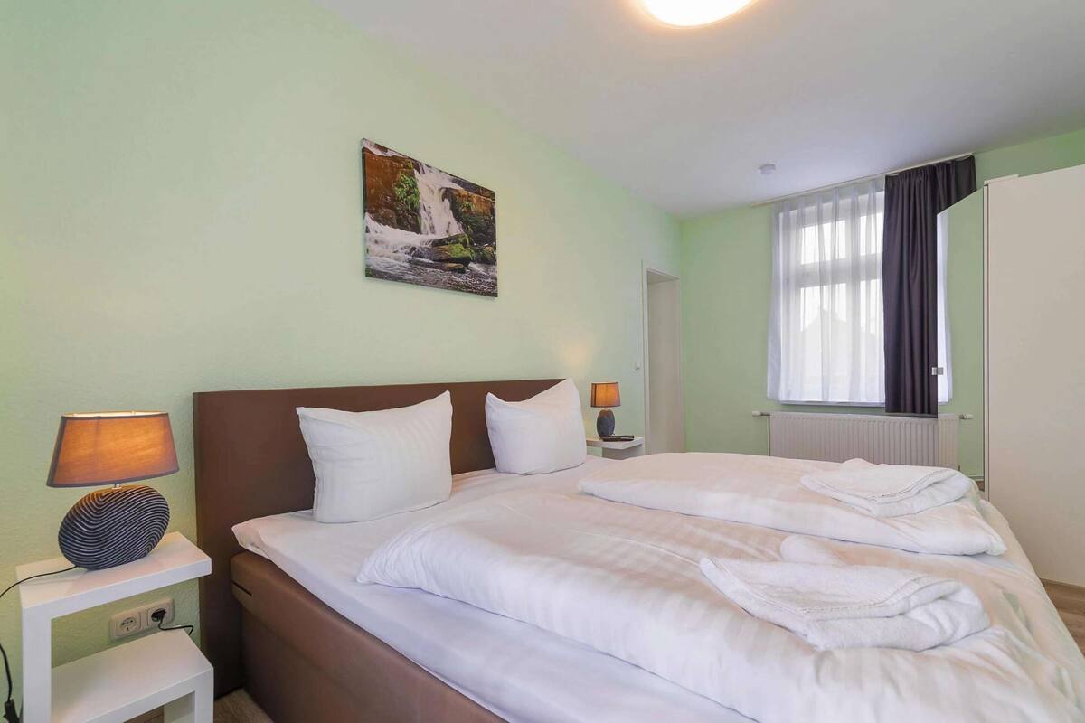 A hotel room with a bed, two bedside lamps, a nightstand, a window with curtains, and a wall-mounted picture.