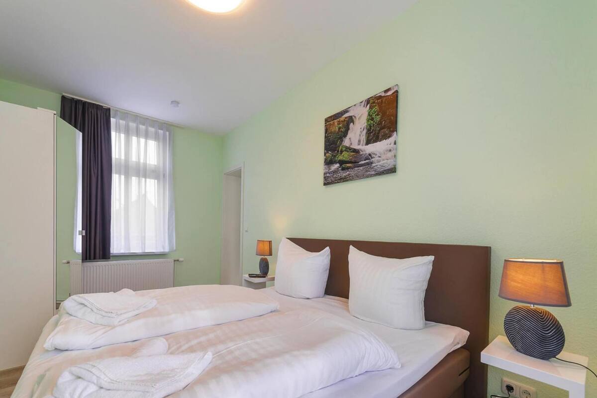 A neatly made bed with white linens and pillows, a bedside table with a lamp, a painting of a waterfall on the wall, and a window with curtains.
