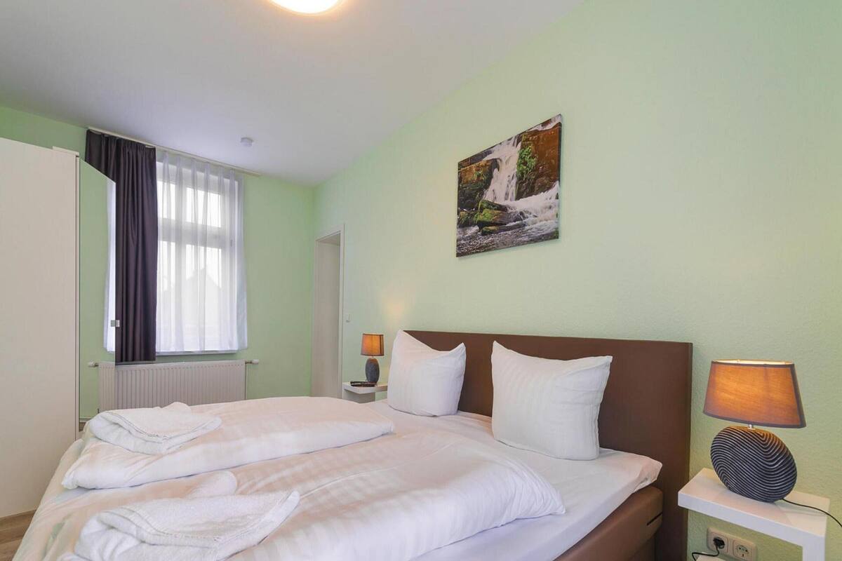 A neatly made bed with white linens and pillows, a bedside lamp, a wall-mounted painting of a waterfall, and a window with curtains.