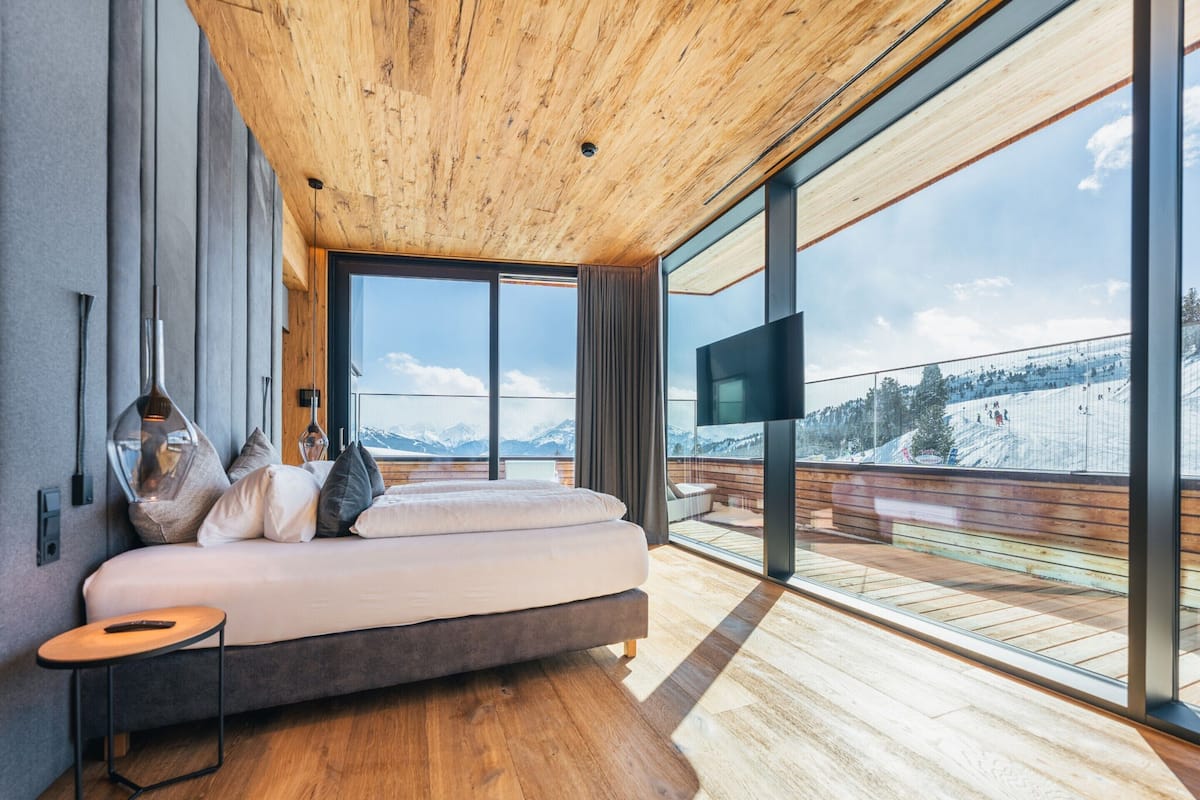 A modern bedroom with a large bed, wooden ceiling, and a view of a snowy mountain landscape.