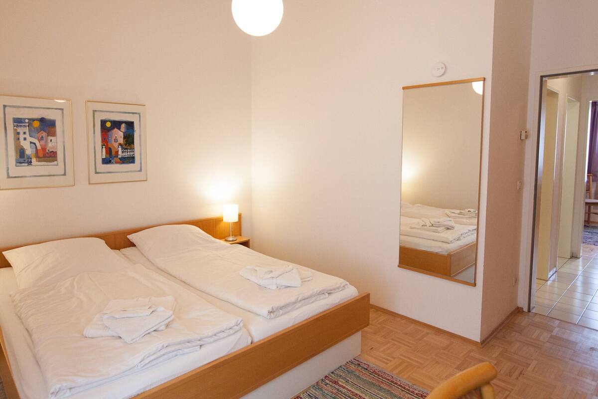 Habitación de hotel con cama doble, mesita de noche con lámpara, espejo y dos cuadros enmarcados en la pared.