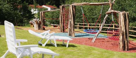 A playground with swings and slides, surrounded by a wooden play structure and red mulch.