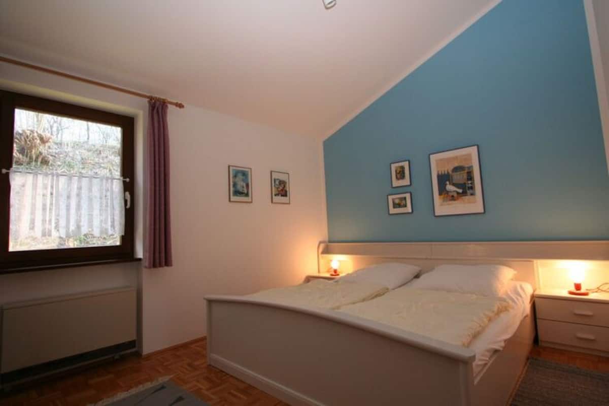 Un dormitorio con cama, mesitas de noche, una ventana con cortinas y cuadros colgados en la pared.