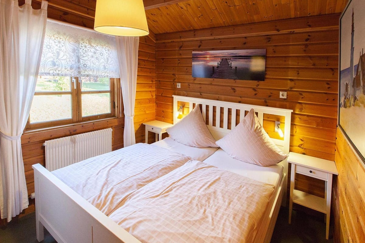 Een houten slaapkamer met een bed, nachtkastjes, een raam met gordijnen en een aan de muur hangend schilderij.