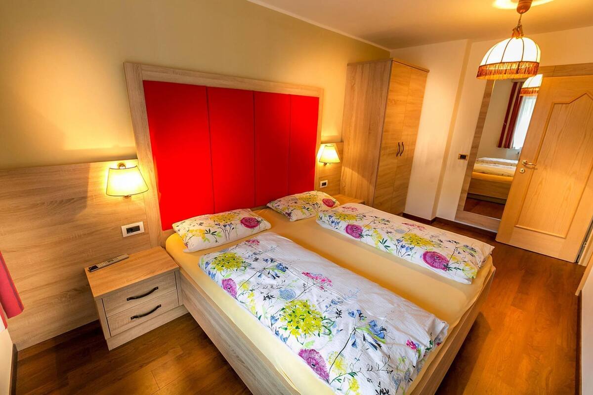A double bed with floral bedding, a red headboard, bedside lamps, a wooden nightstand, and a wardrobe.