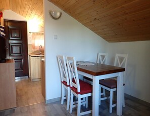 A dining area with a wooden table and white chairs, a kitchen in the background, and a sloped ceiling. - Bungalow in Kolczewo With Parking Space (gmina Wolin)