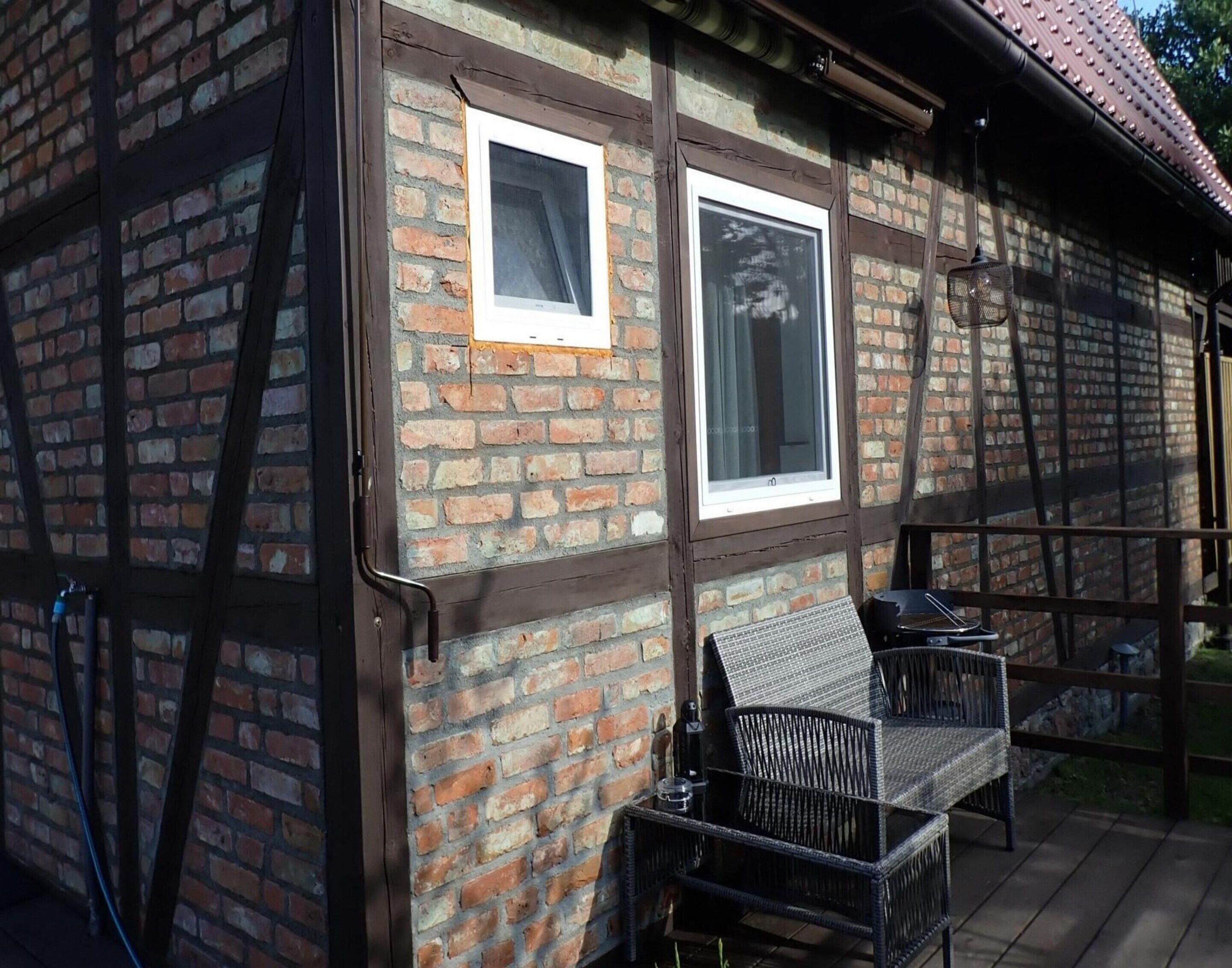 Un bâtiment en briques avec deux fenêtres, une terrasse en bois et du mobilier d’extérieur.