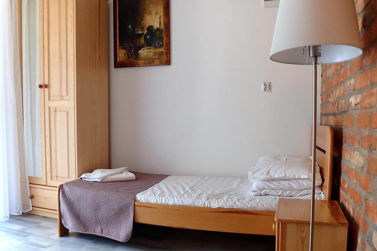 Ein Einzelbett mit weißer Matratze und braunem Kopfteil, ein Holzschrank, ein Bild an der Wand und eine Stehlampe.