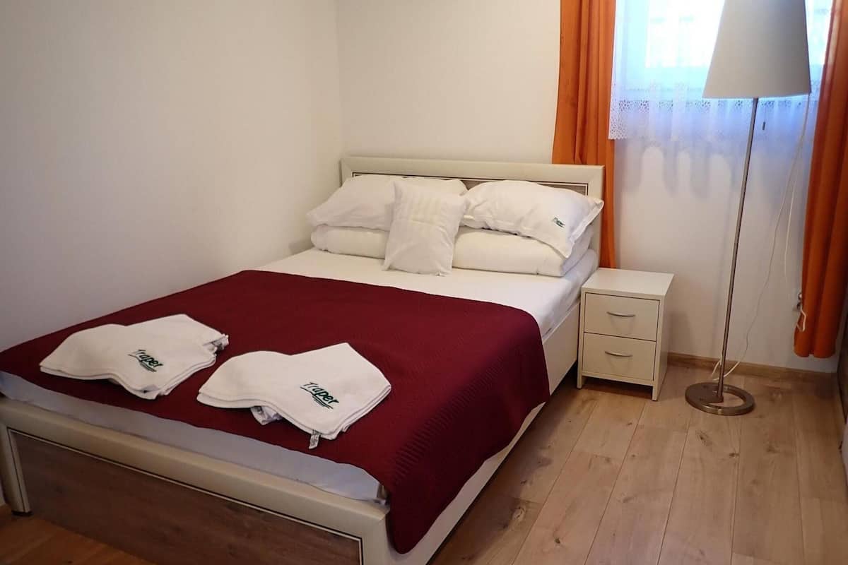 Ein ordentlich bezogenes Bett mit roter Decke und zwei Handtüchern, ein Nachttisch mit Schubladen, eine Stehlampe und ein Fenster mit Vorhängen.