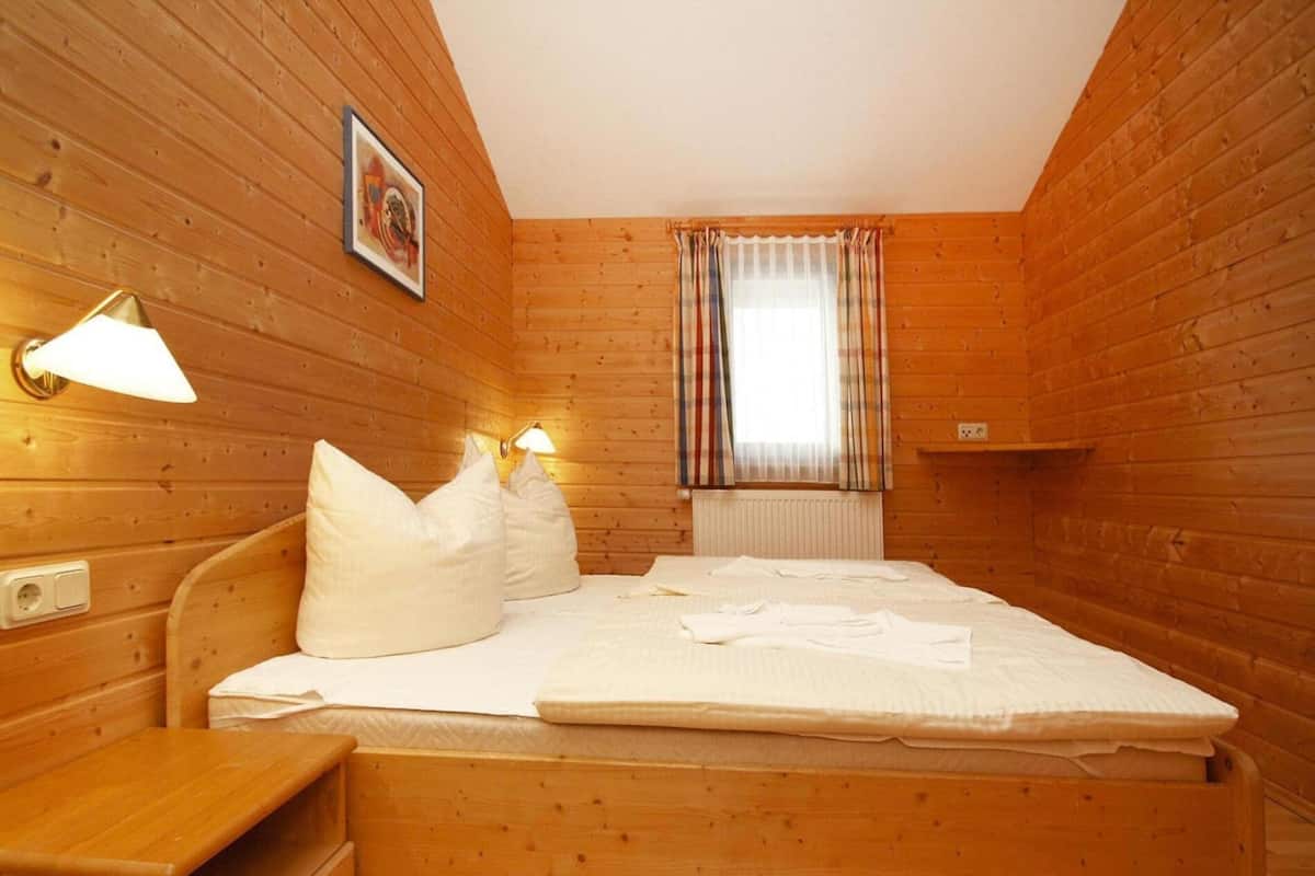 Une pièce chaleureuse, aménagée dans un chalet en bois, avec un lit, des lampes de chevet, une petite table et une fenêtre avec des rideaux.