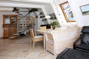 Ein gemütliches Wohnzimmer mit einem Holzesstisch, einem schwarzen Ledersofa und einem Korbstuhl. Eine Treppe mit Holzwandung führt auf eine obere Ebene.