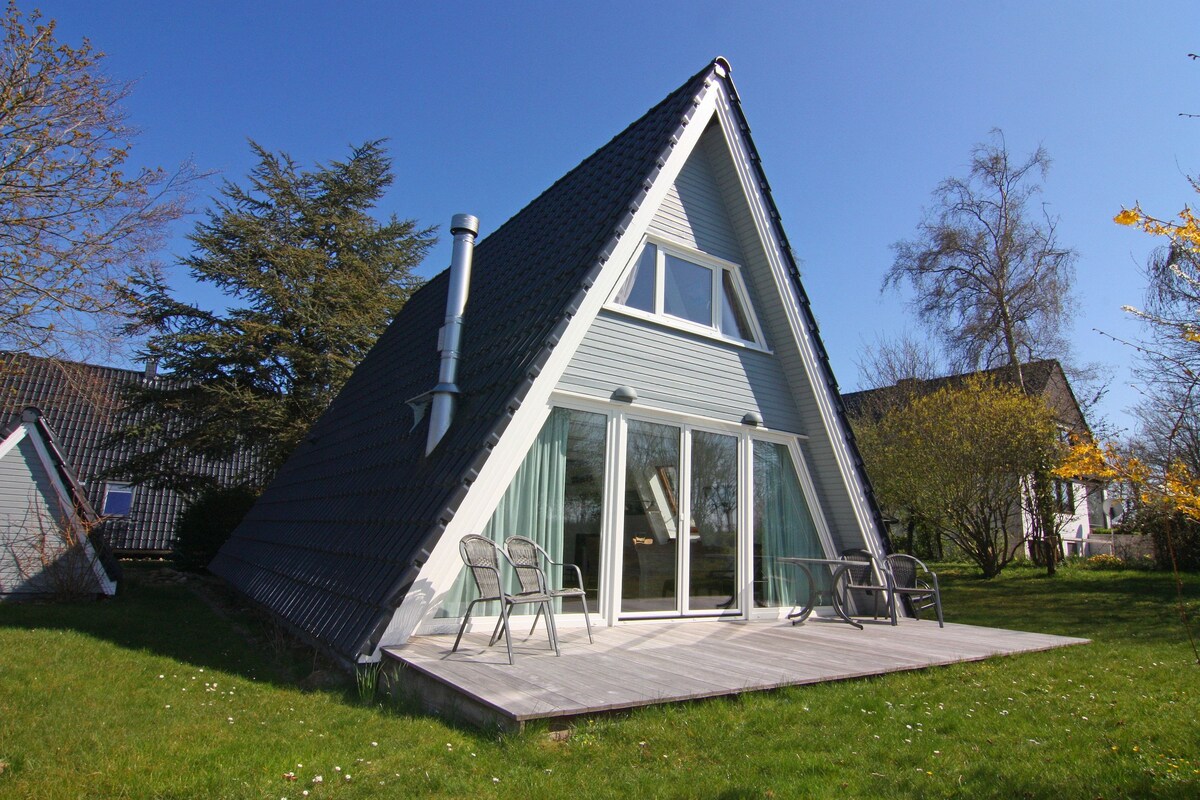 A modern house with a sloped roof, large glass sliding doors, and a wooden deck with chairs.