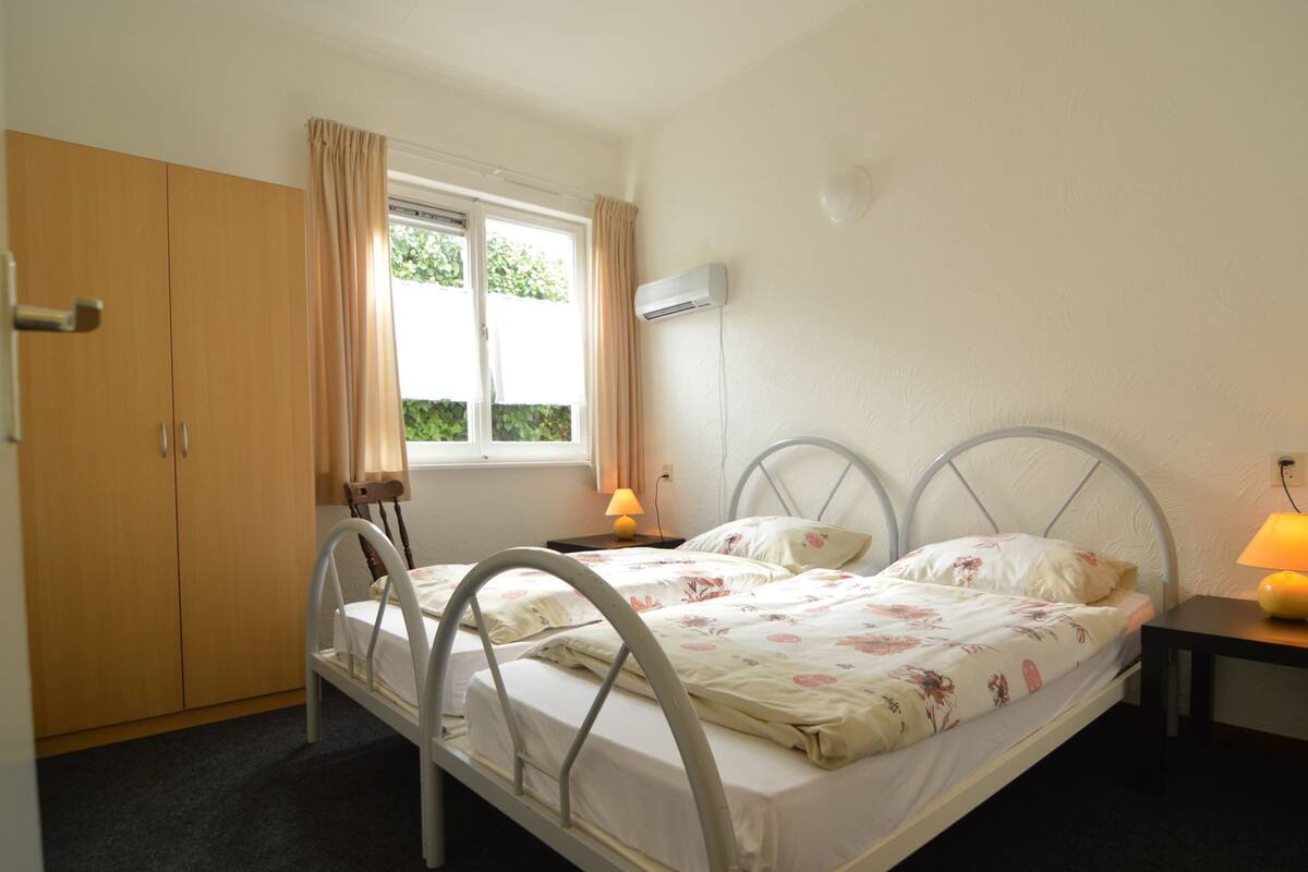Een slaapkamer met twee eenpersoonsbedden, een garderobe, een raam met gordijnen, een bedlampje en een airconditioning aan de muur.