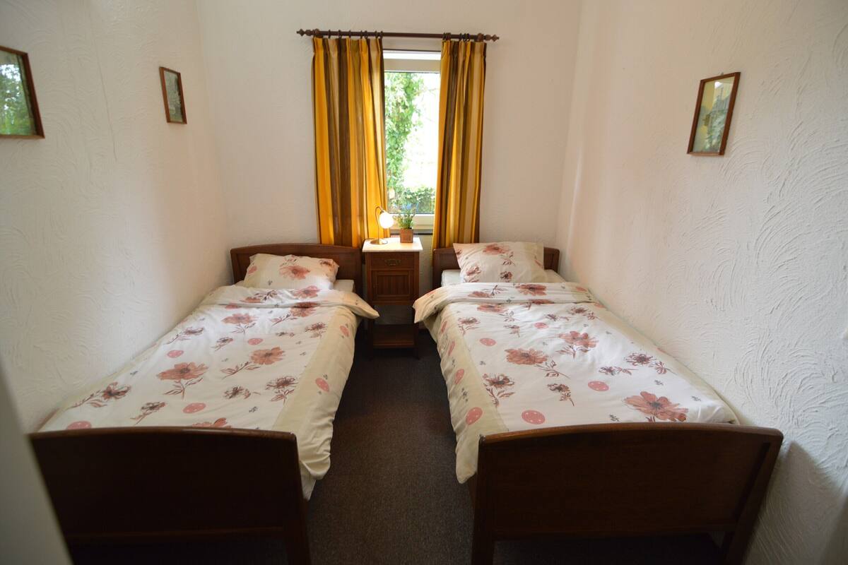 Een kleine kamer met twee eenpersoonsbedden, een nachtkastje met een lamp en een raam met gordijnen.