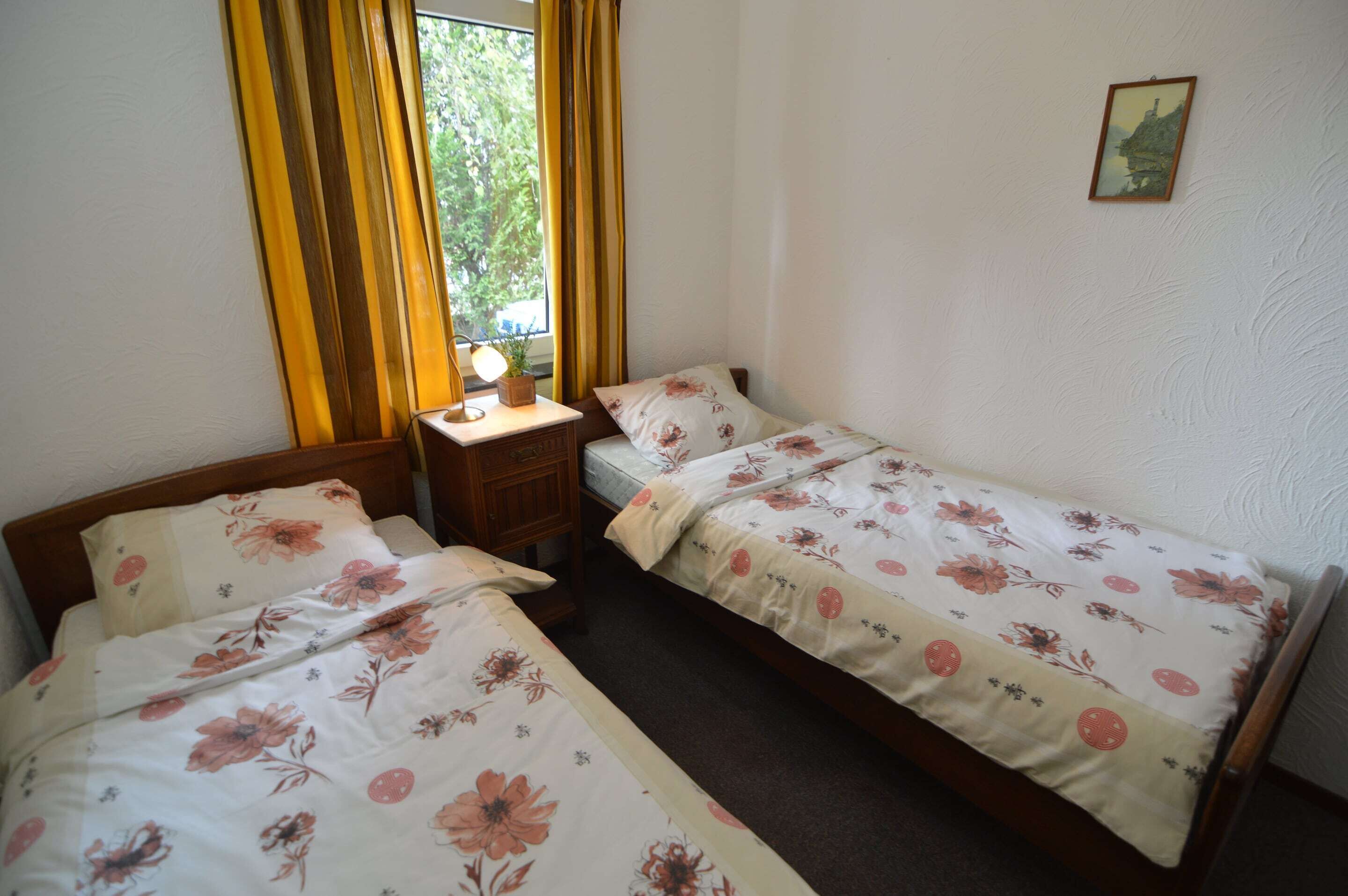 A bedroom with two single beds, a bedside table with a lamp, and a window with yellow curtains.