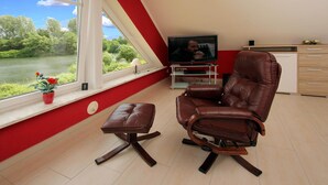 A modern living room with a leather recliner, a small ottoman, a TV on a stand, and a view of a river through a large window. - Apartment House at Kiessee, Westerholt (Nenndorf)