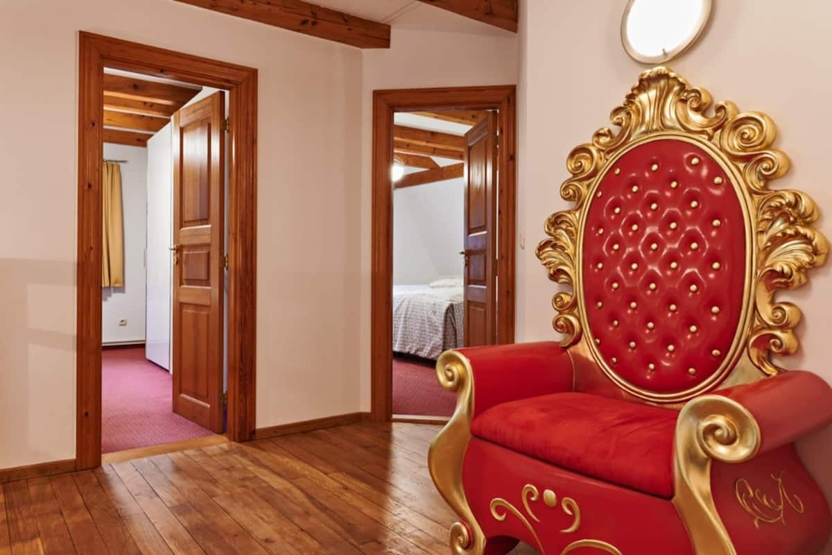 Une pièce dotée d’un parquet en bois, d’un fauteuil rouge recouvert de tissu, agrémenté de détails dorés, et d’un miroir au mur.