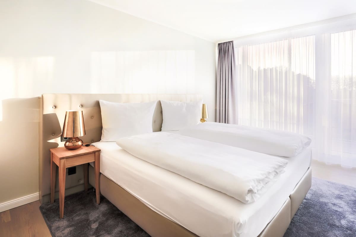 A hotel room with a large bed, a bedside table with a lamp, and a window with sheer curtains.