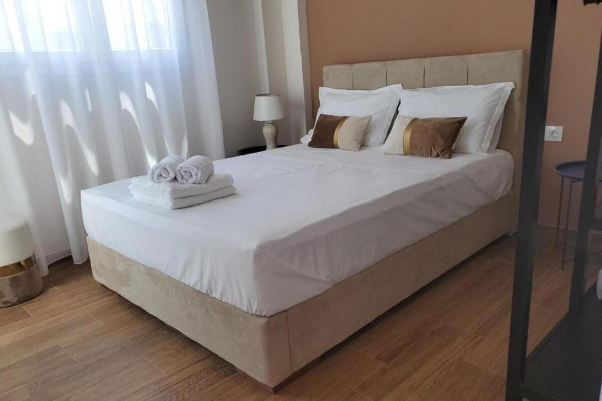 Un lit bien fait, avec du linge de lit blanc et une tête de lit beige, une lampe de chevet et une petite table ronde placée à droite.
