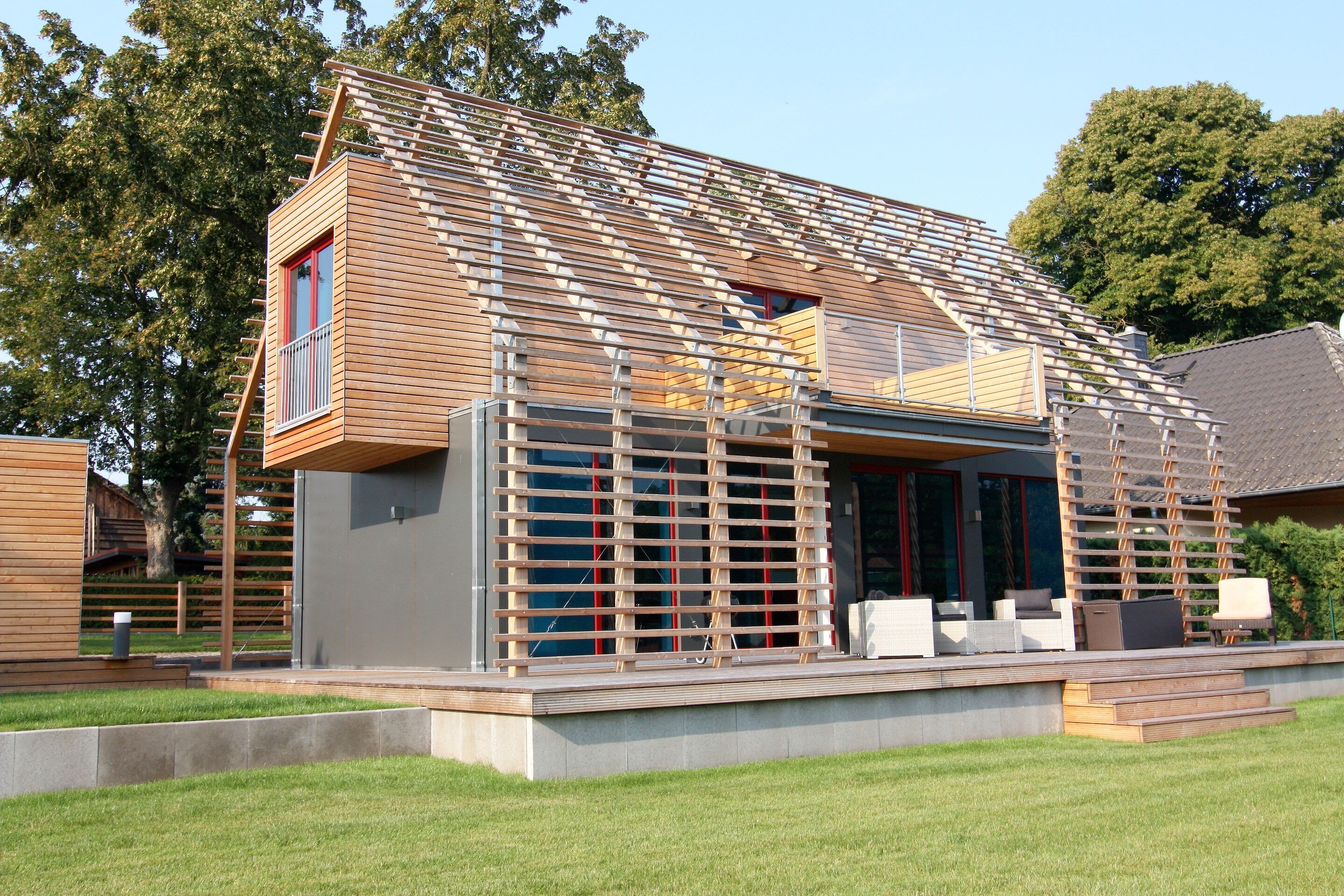 Una casa moderna con techo inclinado, fachada de madera y balcón con mobiliario exterior.