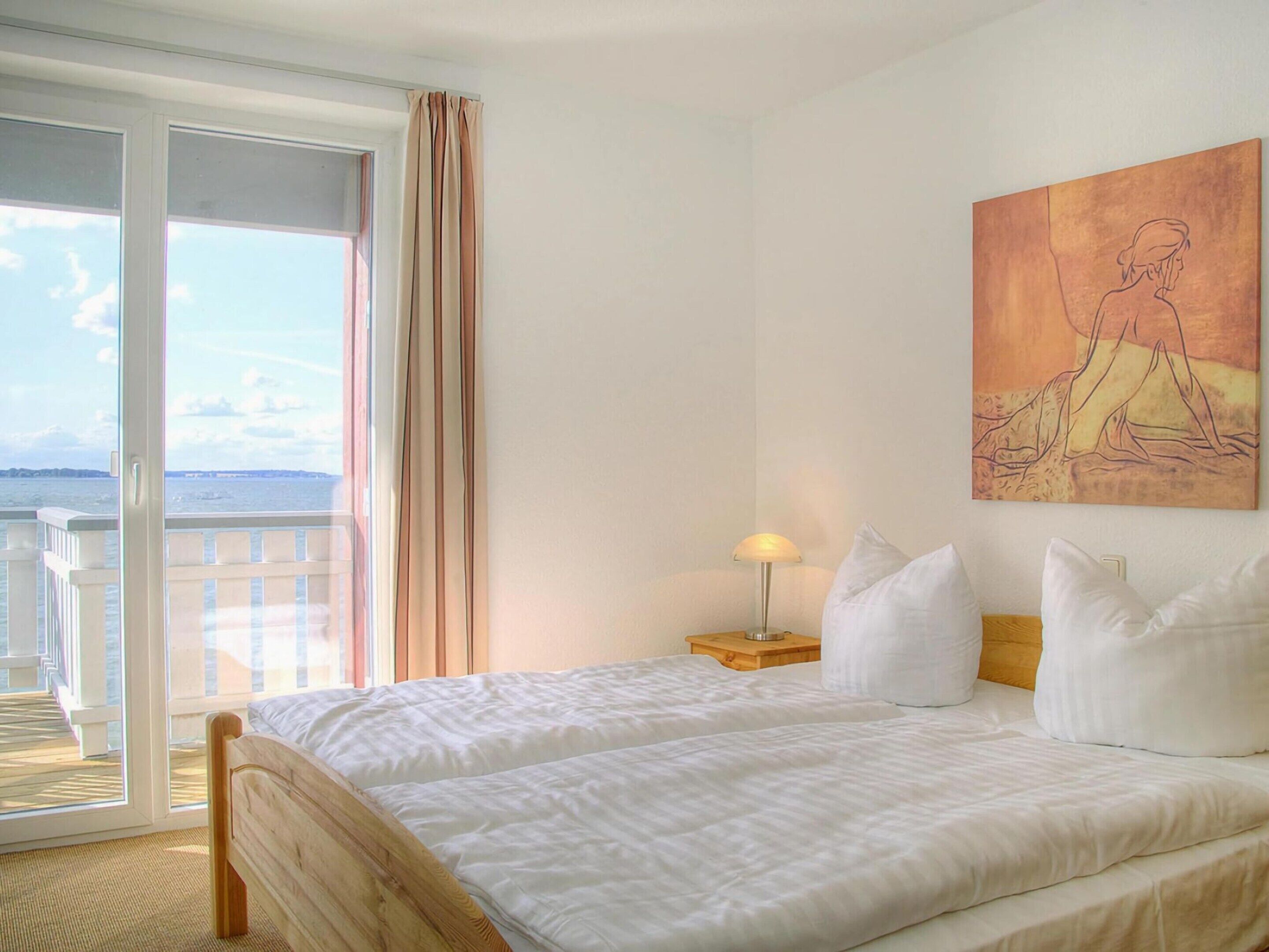 A hotel room with a bed, a nightstand, a lamp, a painting, and a view of the sea through a sliding door.