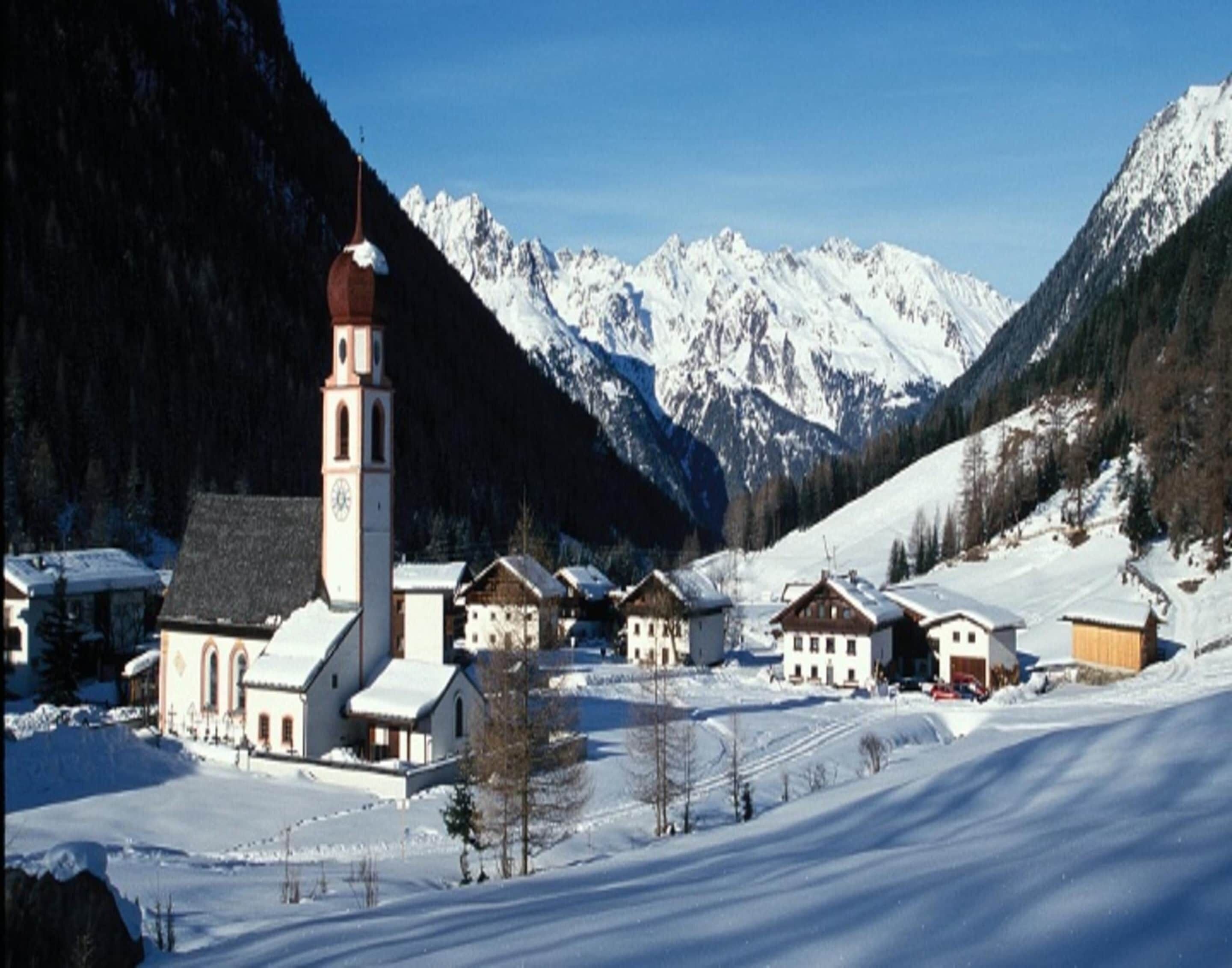 Ein verschneites Dorf mit einer Kirche und mehreren traditionellen Häusern, umgeben von Bergen.