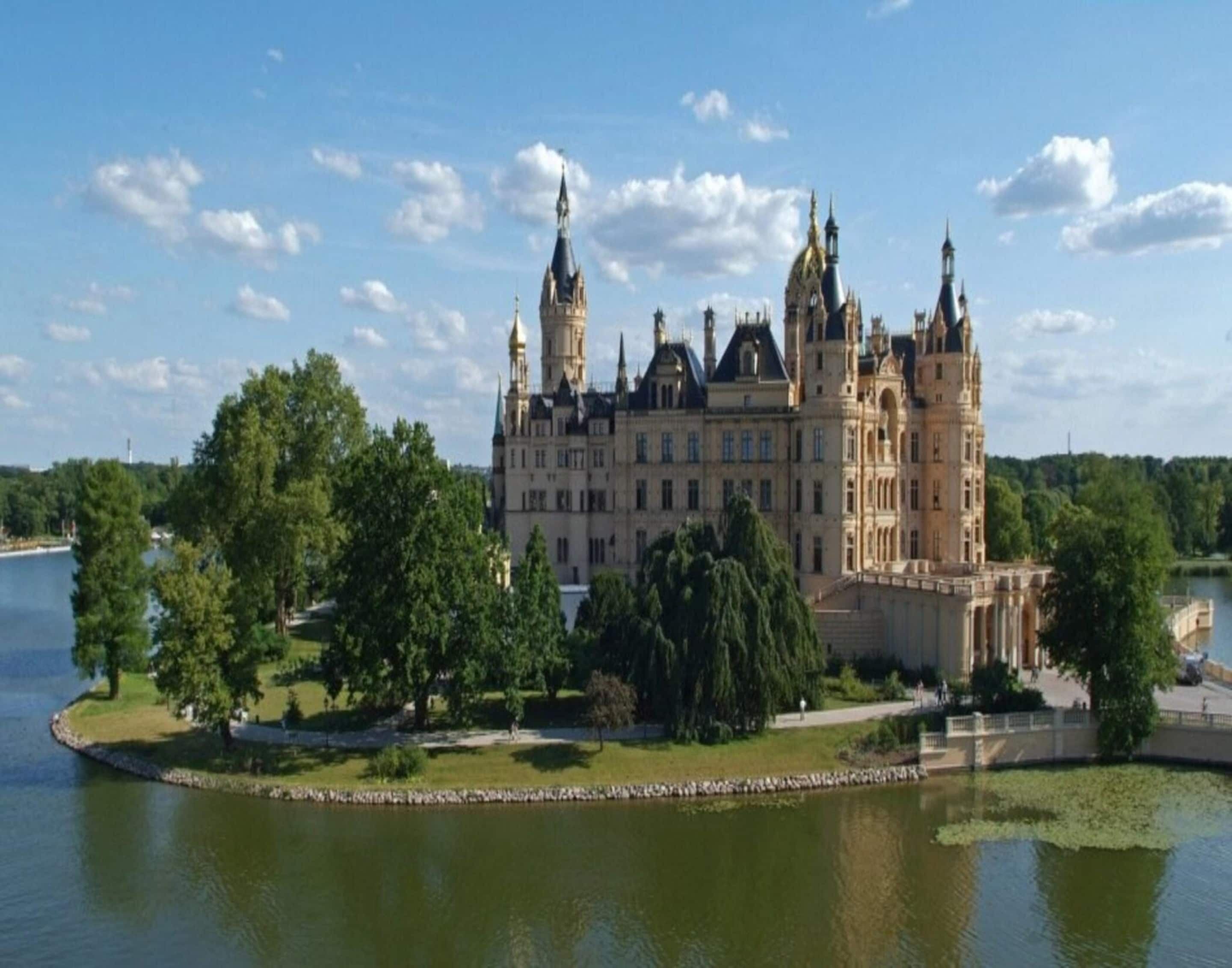 A large, historic castle with multiple towers and a moat, surrounded by lush greenery and a body of water.
