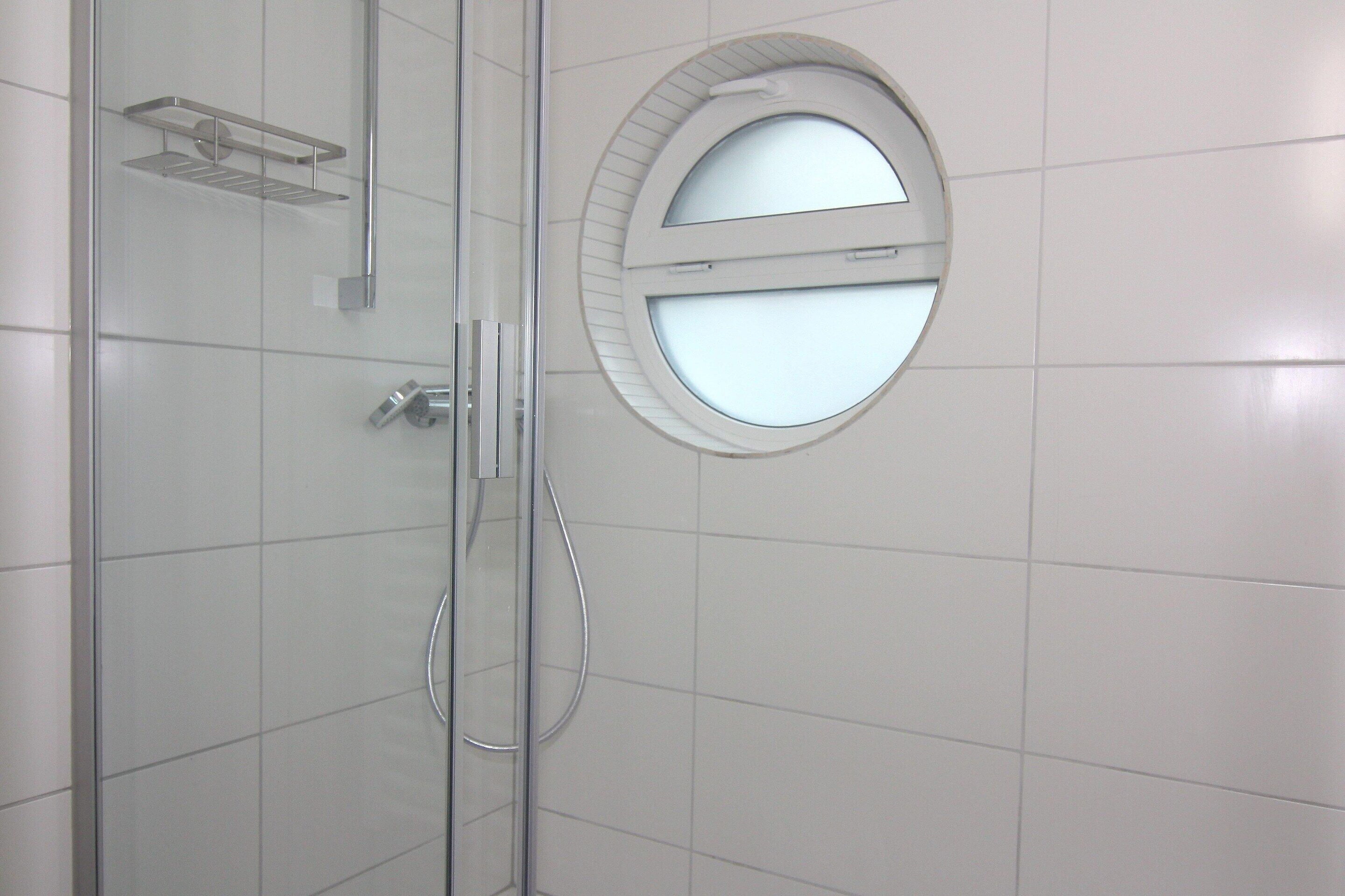 A modern bathroom with white tiles, a glass shower enclosure, a round mirror with a door, and a metal shelf.