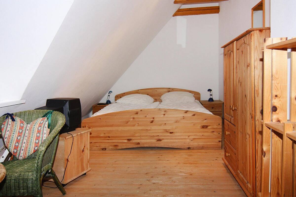 Dormitorio tipo loft con techo inclinado, suelo de madera, cama con ropa de cama blanca, una silla de mimbre verde y un armario de madera.