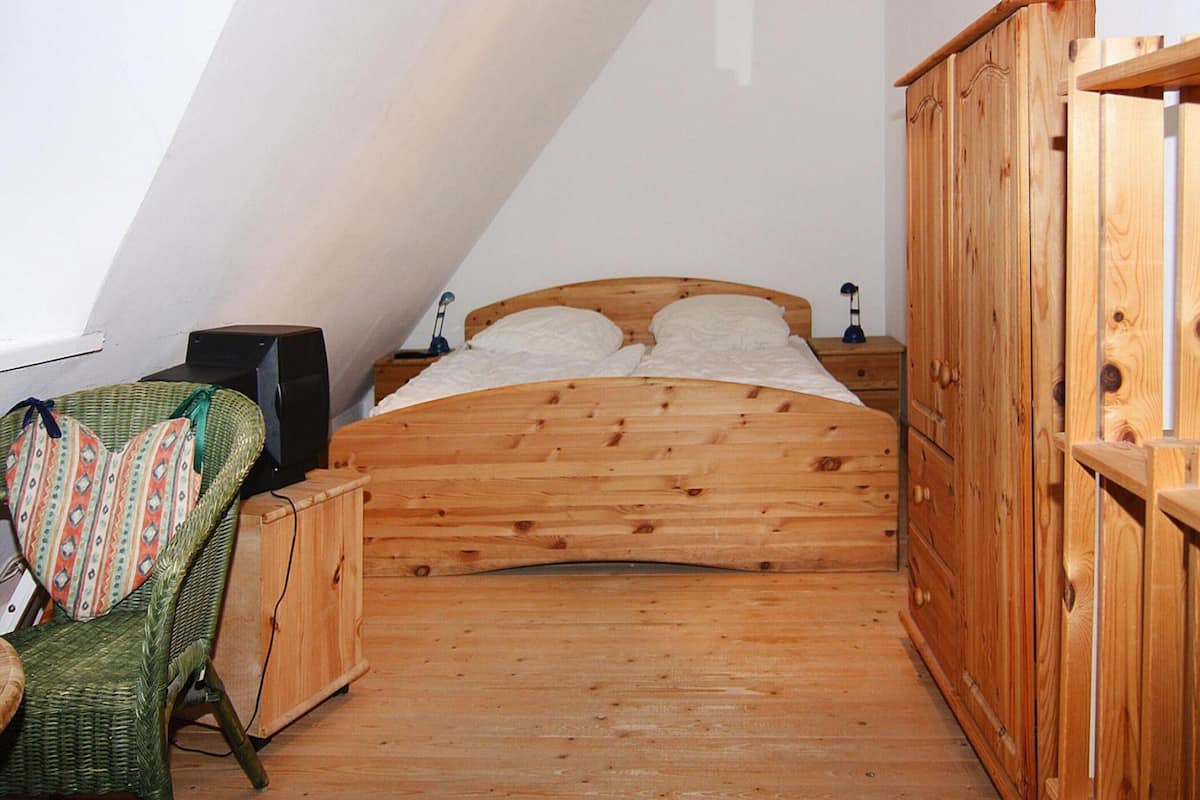 Una habitación pequeña con estructura de madera, techo inclinado, una cama individual, una silla de mimbre verde y un armario de madera.