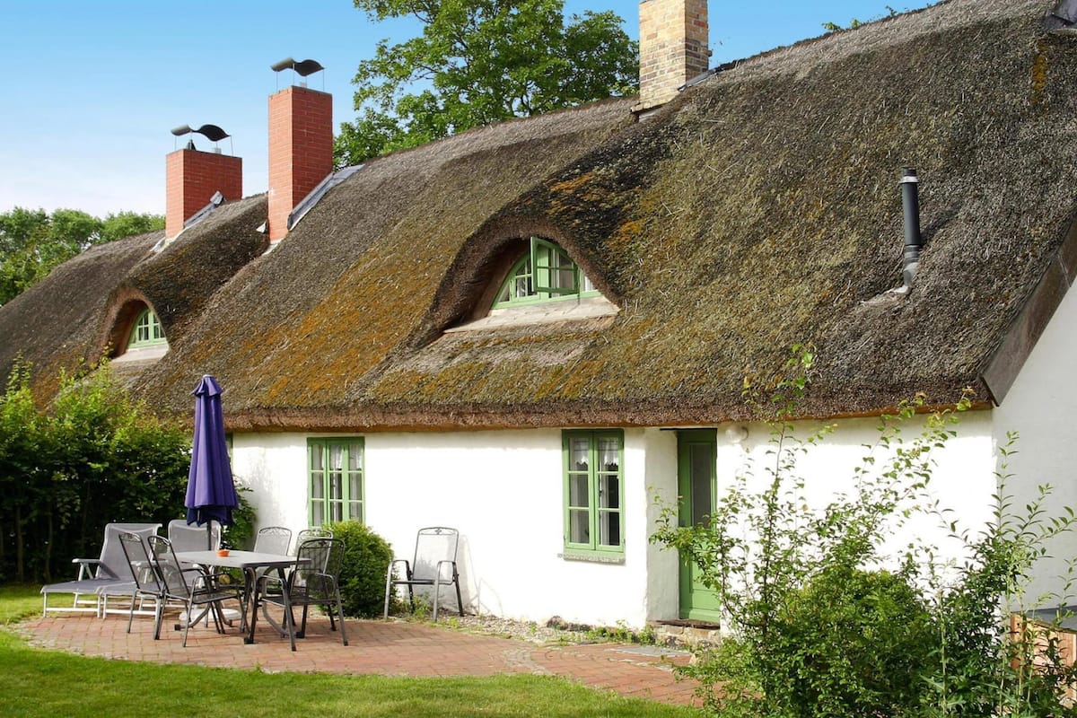 A traditional thatched-roof cottage with a small patio area, a table with chairs, and a blue umbrella.