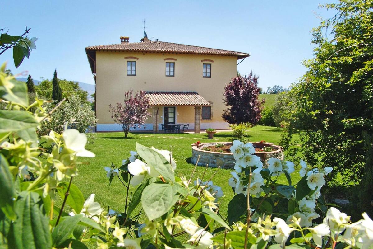 Una casa a due piani con tetto in tegole, un portico e un giardino curato con piante fiorite e un aiuola circolare.