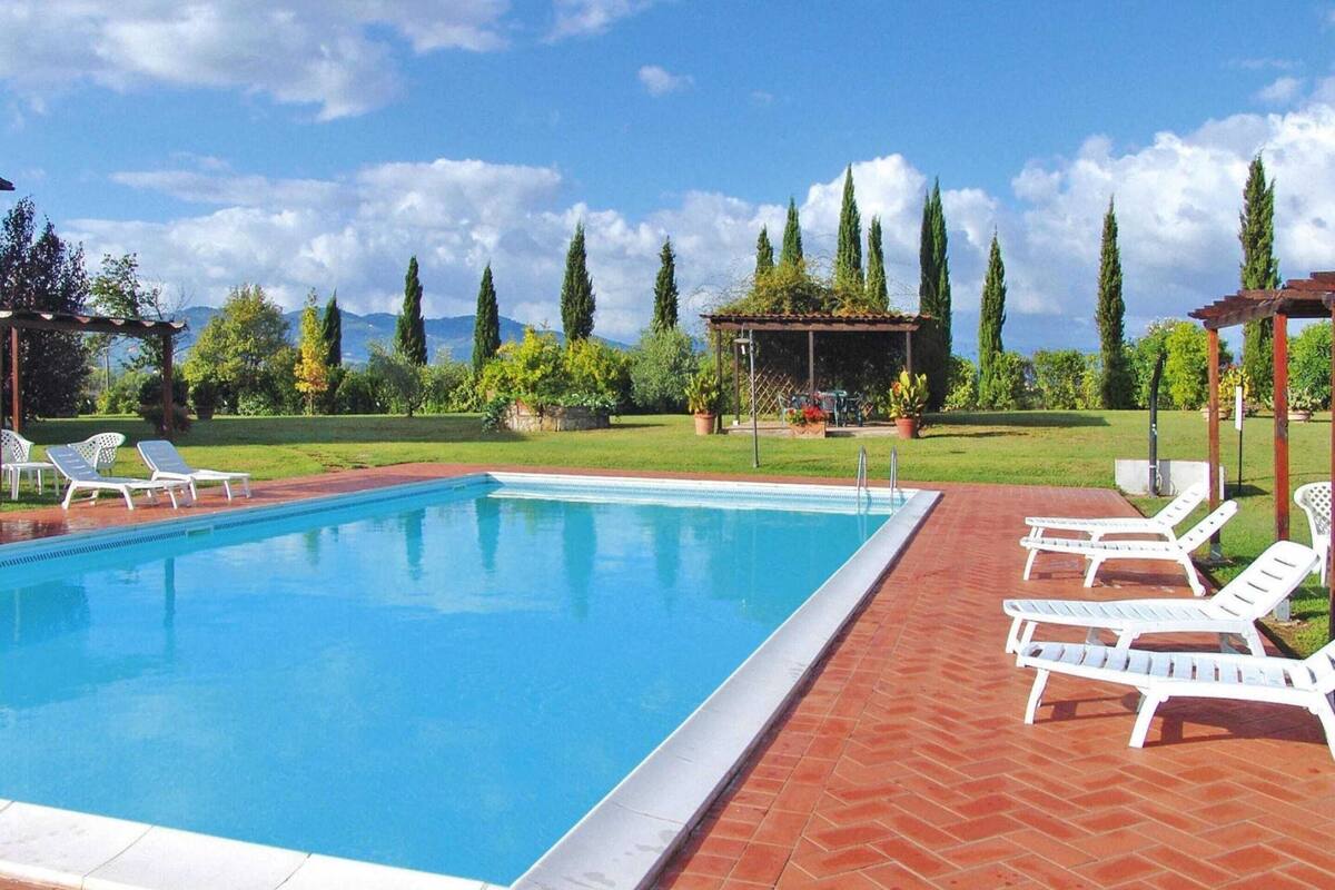 Una piscina rectangular con borde de azulejos, rodeada por un patio de ladrillo. Se ven sillas de descanso blancas alineadas junto a la piscina.