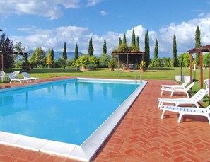 Una piscina rectangular con borde alicatado, rodeada por un patio de ladrillo. Se observan sillas reclinables blancas y una estructura de pérgola al fondo.