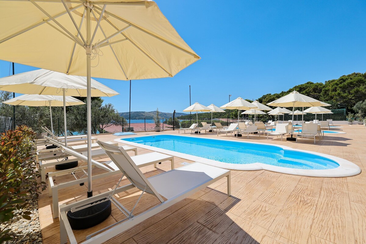 Un espace aménagé au bord de la piscine, avec des chaises longues et des parasols, un ciel d’un bleu éclatant et une vue sur la mer.