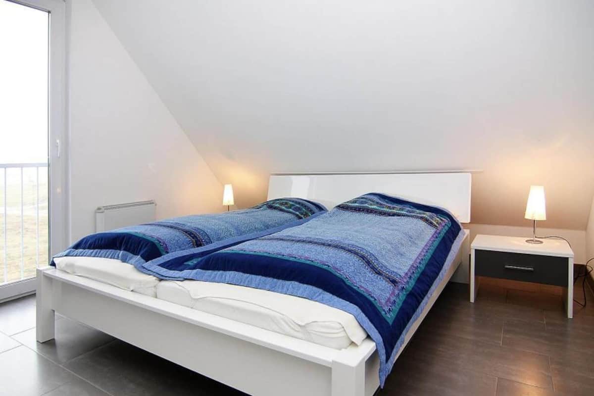 Una camera da letto moderna con un letto bianco, due comodini con lampade e un'ampia finestra che lascia entrare luce naturale.
