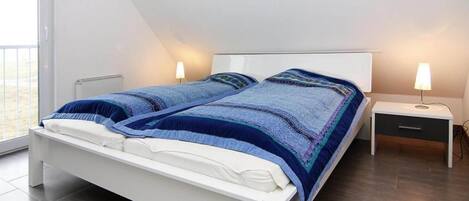 Un dormitorio moderno con una cama blanca, dos mesitas de noche con lámparas y un amplio ventanal que proporciona luz natural.