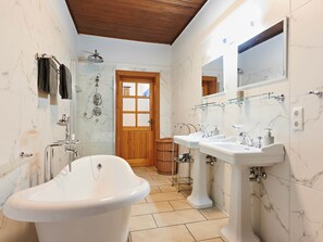 Ein Badezimmer mit freistehender Badewanne, zwei Waschbecken auf Säulen, einer Dusche mit Handbrause und einer Holztür.