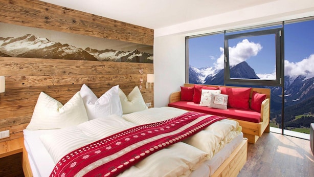 A bedroom with a wooden headboard, a large bed with white bedding, a red sofa, and a mountain view through large windows. - Apartment in Neukirchen With a Balcony (Neukirchen am Grossvenediger)