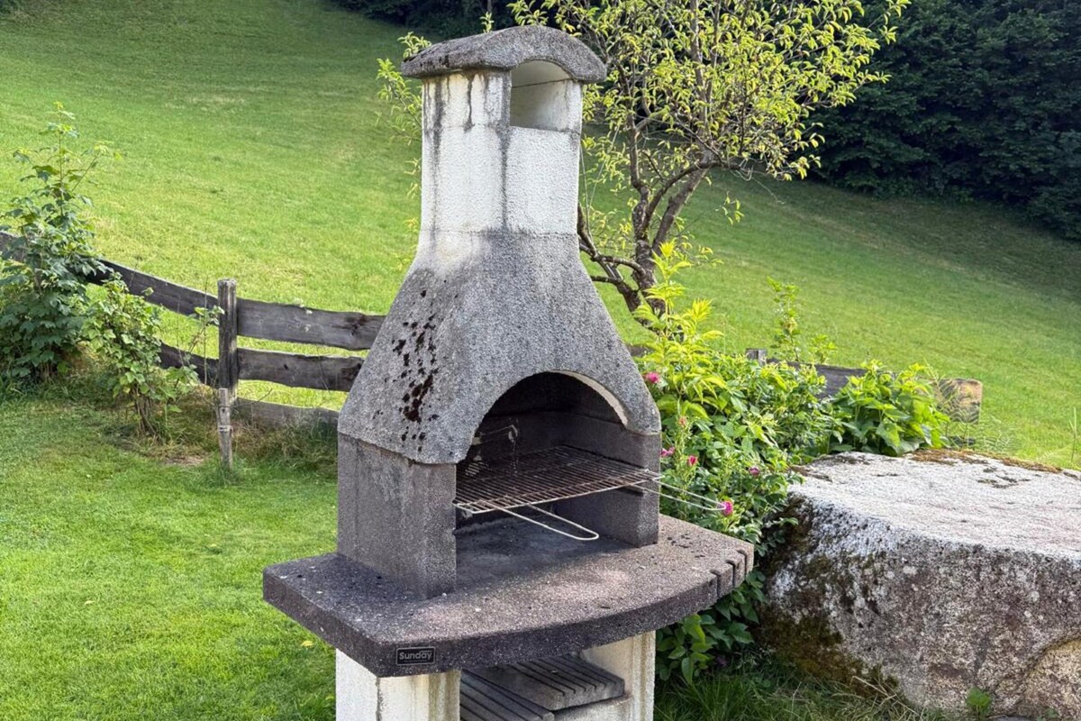 Un four extérieur en pierre, doté d’une grille en métal, entouré d’une zone herbeuse et d’une clôture en bois.