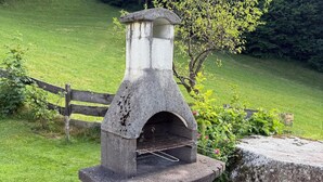 A stone-built outdoor oven with a metal grill, set against a grassy field with a wooden fence and trees in the background. - Apartment in Neukirchen With a Balcony (Neukirchen am Grossvenediger)