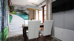 Un coin repas agrémenté d’une fresque représentant un sentier en bois menant à un plan d’eau, avec des chaises blanches, une table et un téléviseur fixé au mur.