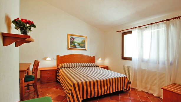 Una camera da letto con un letto, una sedia, un tavolo, uno scaffale con fiori e un quadro appeso al muro, oltre a una finestra con tende.