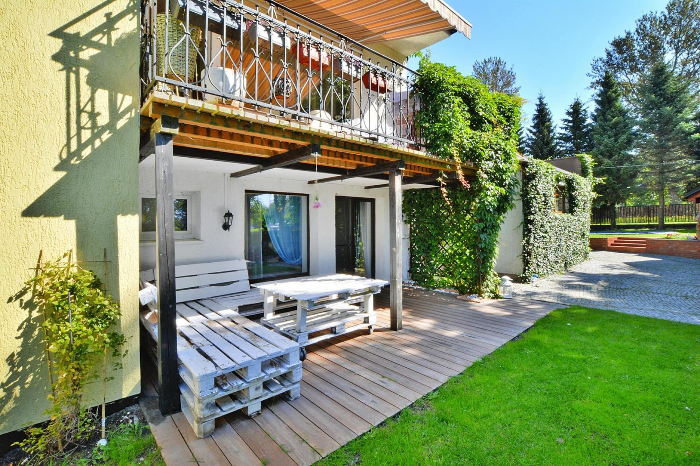 Une terrasse en bois avec une table et des bancs, un espace extérieur couvert avec une rambarde, et un jardin entouré d’une clôture en bois.