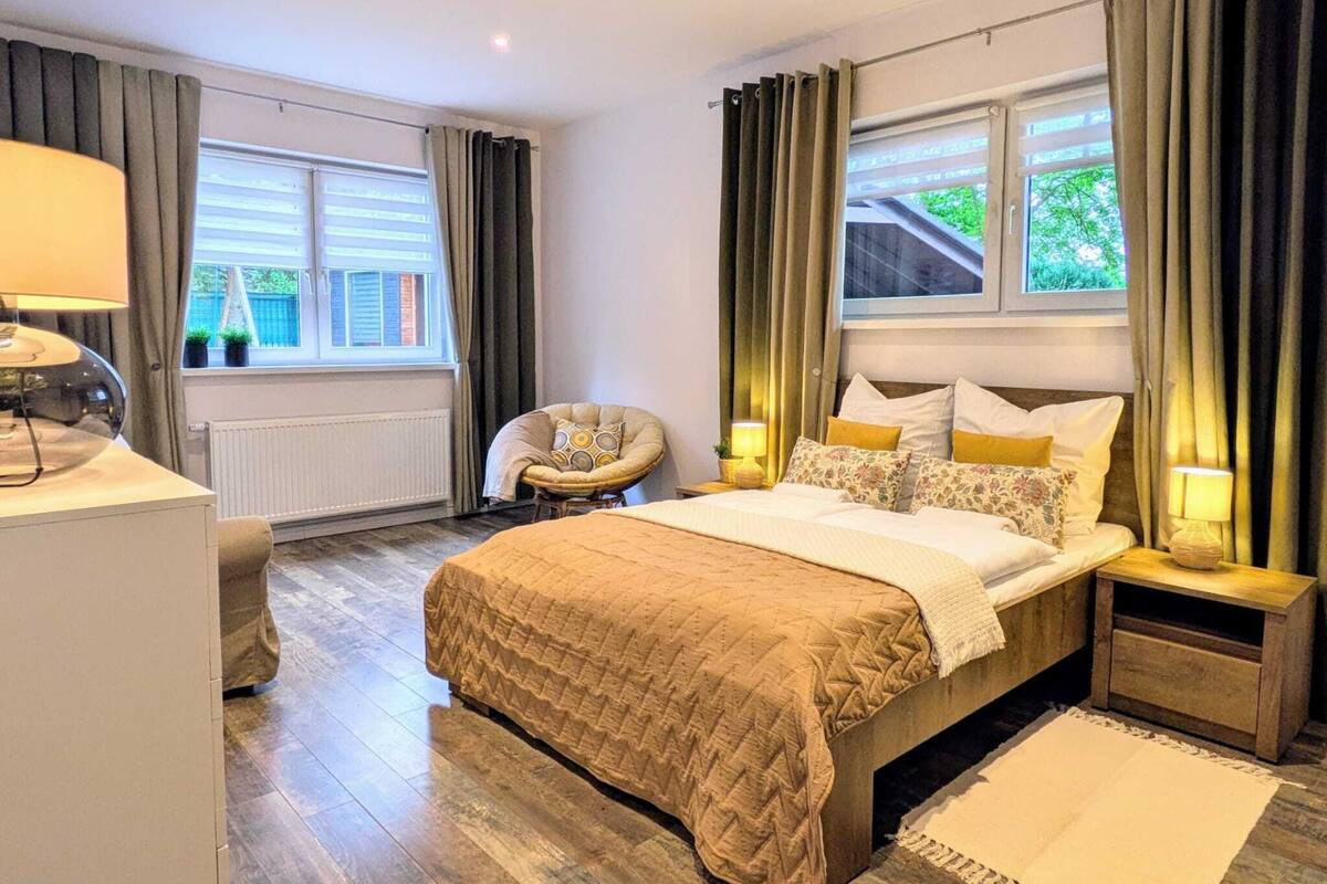 A modern bedroom with a large bed, bedside lamps, a chair, and a view of greenery outside.