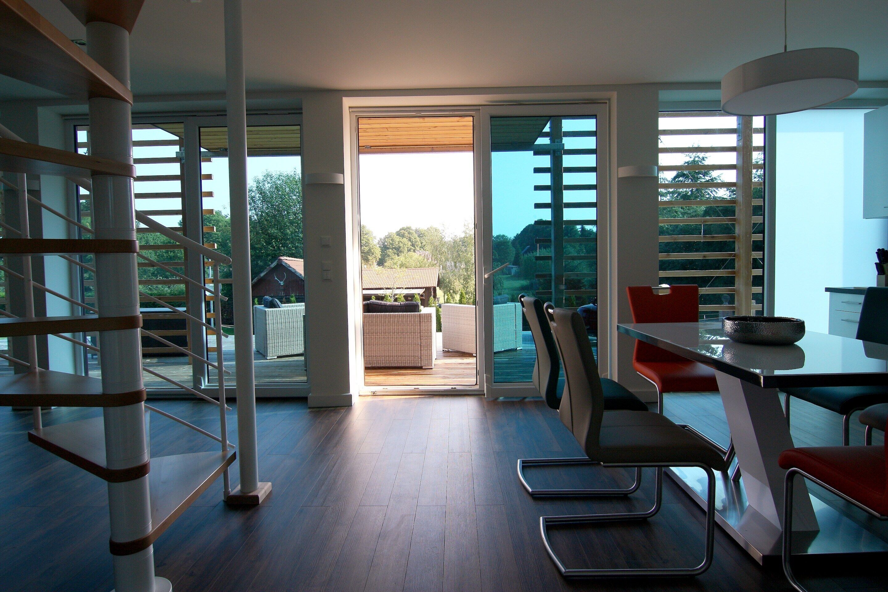 A modern interior with a staircase, a dining area with chairs, and a view through a glass door to an outdoor seating area.