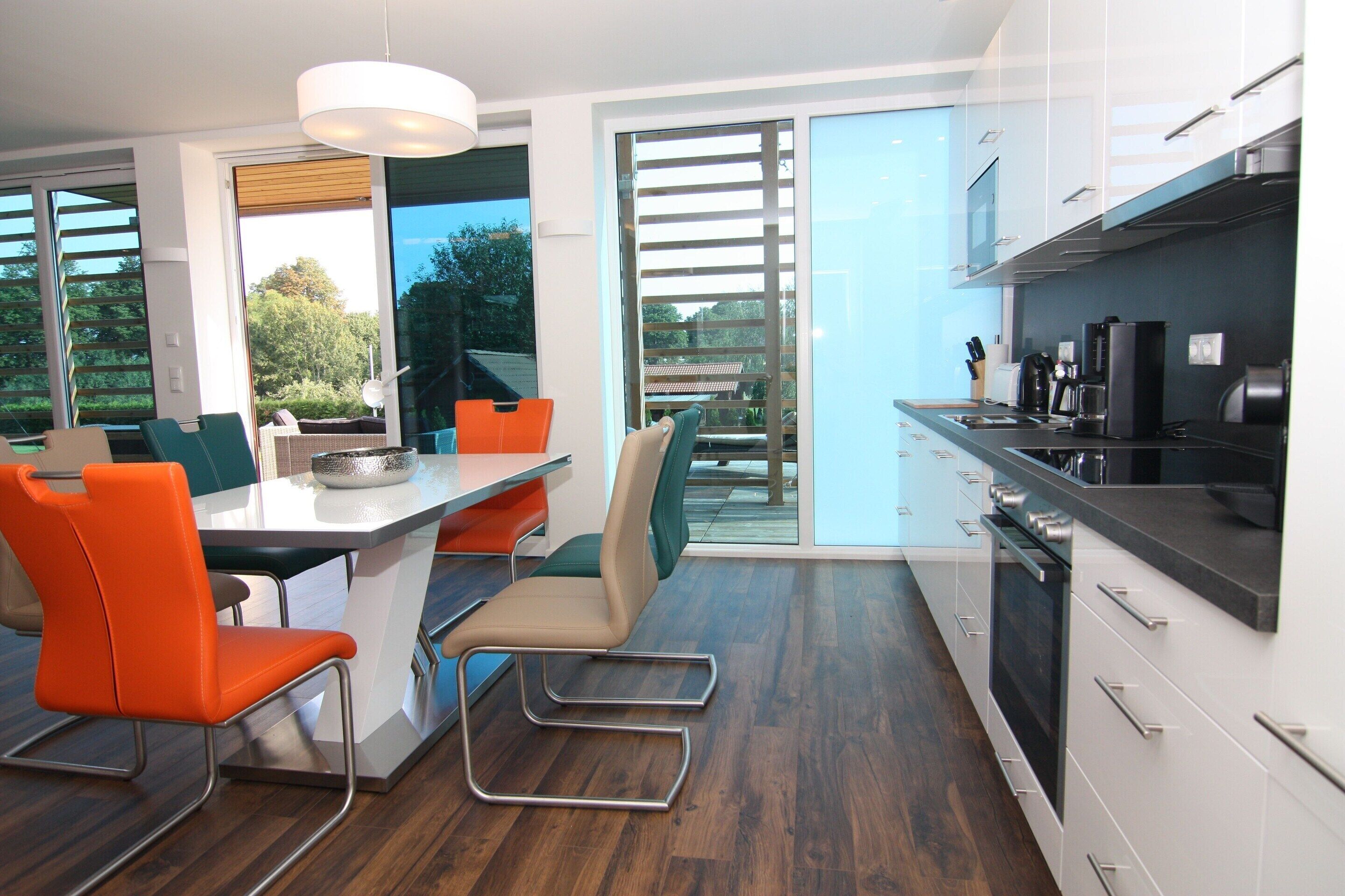 A modern kitchen with white cabinets, a black countertop, and a built-in oven. There is a dining area with orange chairs and a table.
