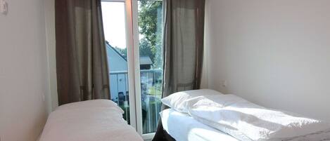 A small, clean room with two single beds, a sliding glass door leading to a balcony, and a view of the outside.