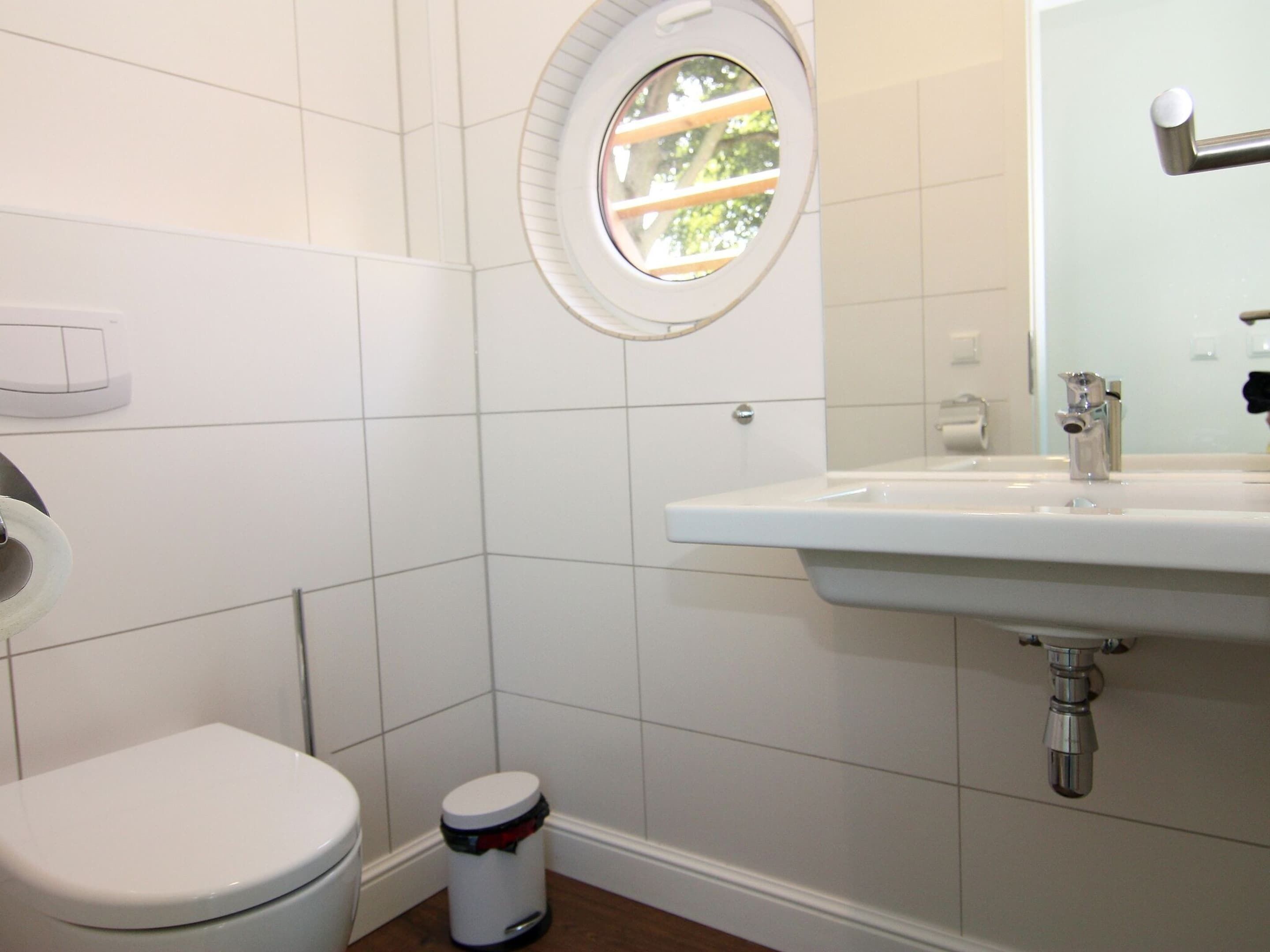 A bathroom with a toilet, a sink, a trash bin, and a window with a view of trees.