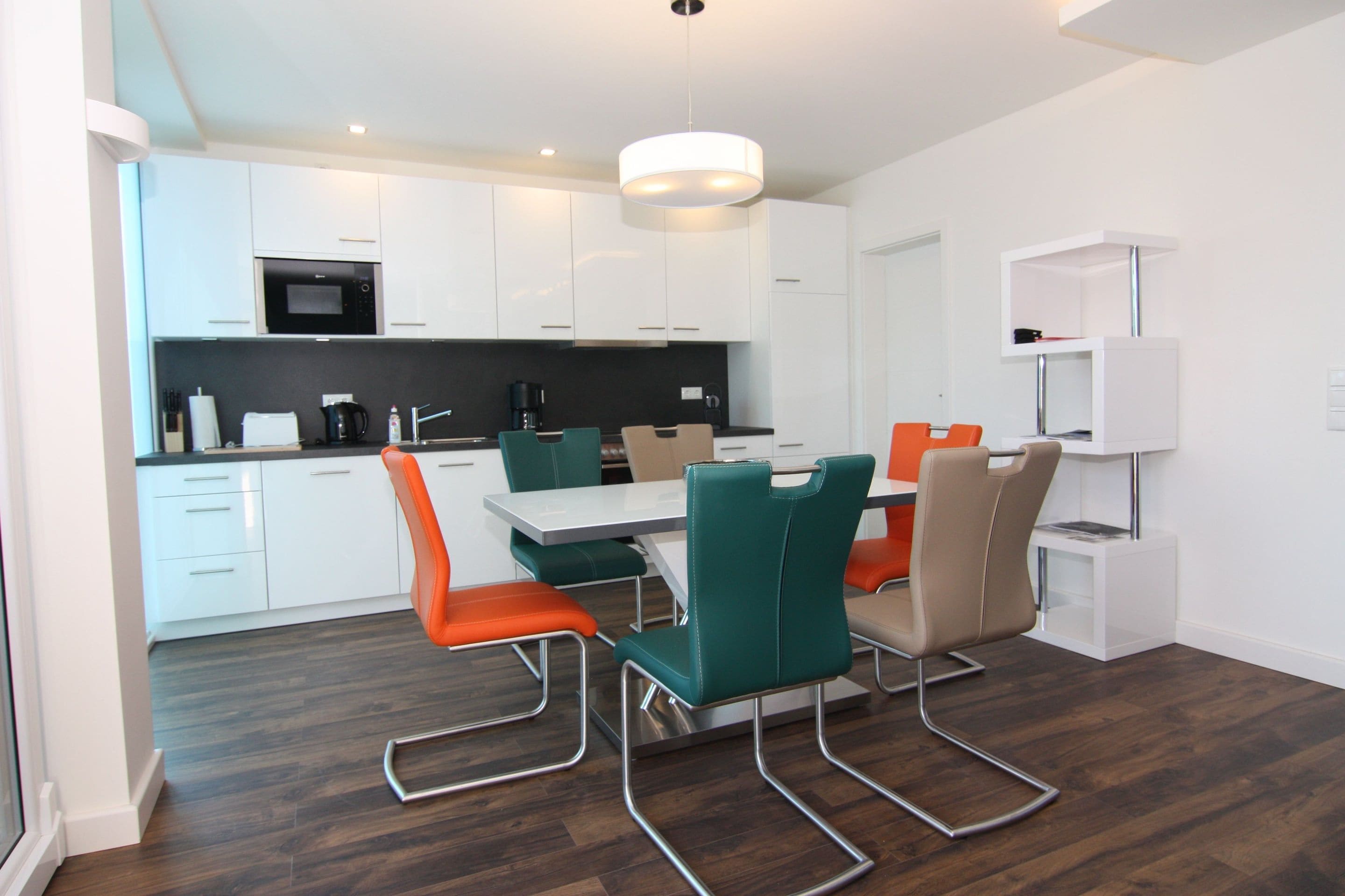 A modern kitchen with white cabinets, a black countertop, and a dining area with a white table and orange chairs.